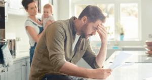 (man stressing over bills while his wife holds their baby) 