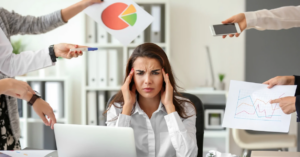 (Stressed woman at a desk surrounded by charts, phones, and hands pointing, representing the pressure and overwhelm caused by financial stress and debt.) 