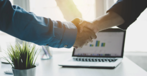 (Two people shaking hands in front of a laptop displaying charts, with sunlight streaming in and a small plant on the desk.)