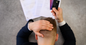 A man sitting at a table with his head in one hand and credit cards in the other, with bills and paperwork spread out in front of him.