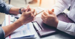Close-up of two people sitting at a table with their hands clasped, surrounded by financial documents, a smartphone, and a leather portfolio. 