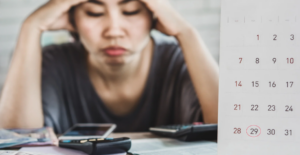 (A woman sitting with her head in her hands, looking stressed, with a calendar showing the 29th circled and financial documents and a calculator on the table.)