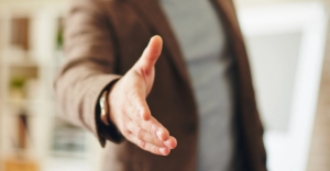 (A close-up of a person extending their hand for a handshake, wearing a brown jacket with a blurred indoor background.)