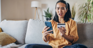 (A woman sitting on a couch looking shocked while holding a credit card in one hand and a smartphone in the other, with a concerned expression on her face.)